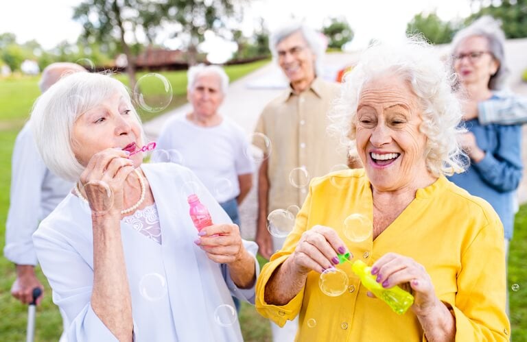 Two Elderly Women Blowing Bubbles Joyfully with Friends Outdoors at an Independent Living Community