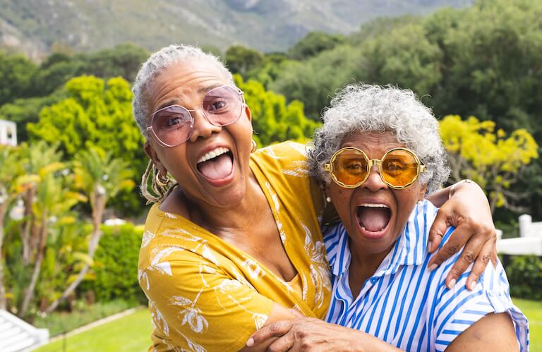 Two Joyful Elderly Women Laughing and Embracing Outdoors Wearing Sunglasses at an Independent Living Facility